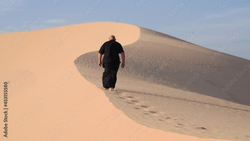 Old man in black is engaged in kung fu in the dunes at sunset. a lot of sand, golden sunset light. sports training. footprints in the sand. Man is going higher and higher to the desert hill. 