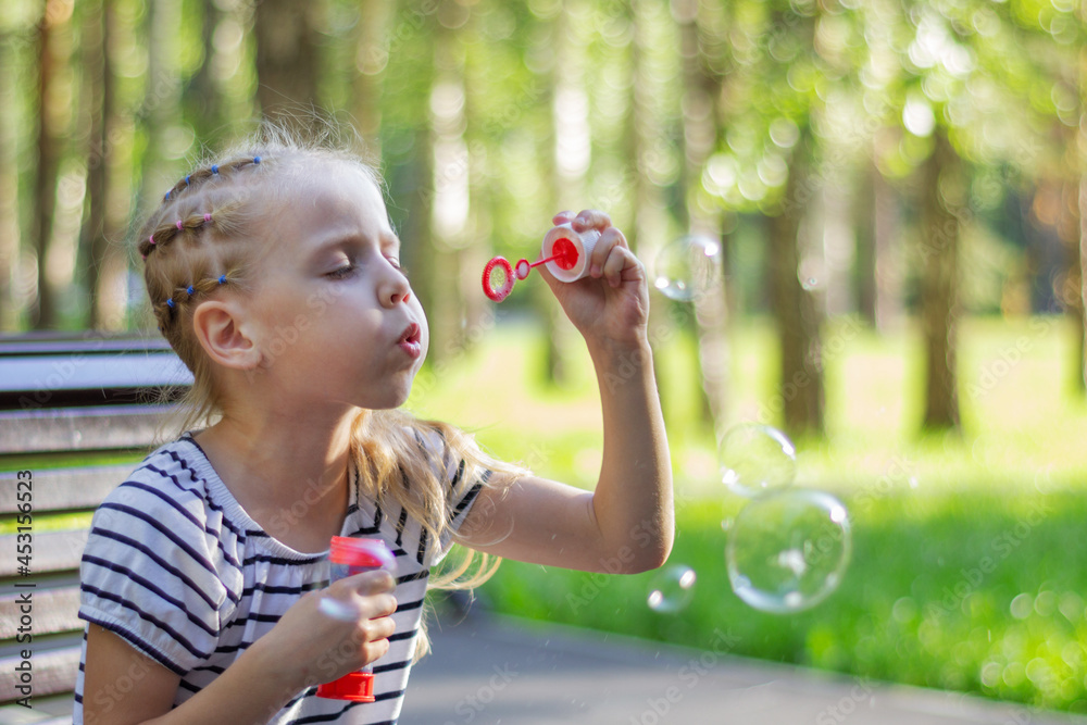 A little blonde girl in a striped T-shirt on vacation sits on a park bench in good weather and happily blows soap rainbow bubbles close-up. There is a lot of sunshine and green trees around. A little blonde girl in a striped T-shirt on vacation sits on a park bench in good weather and happily blows soap rainbow bubbles close-up. There is a lot of sunshine and green trees around.