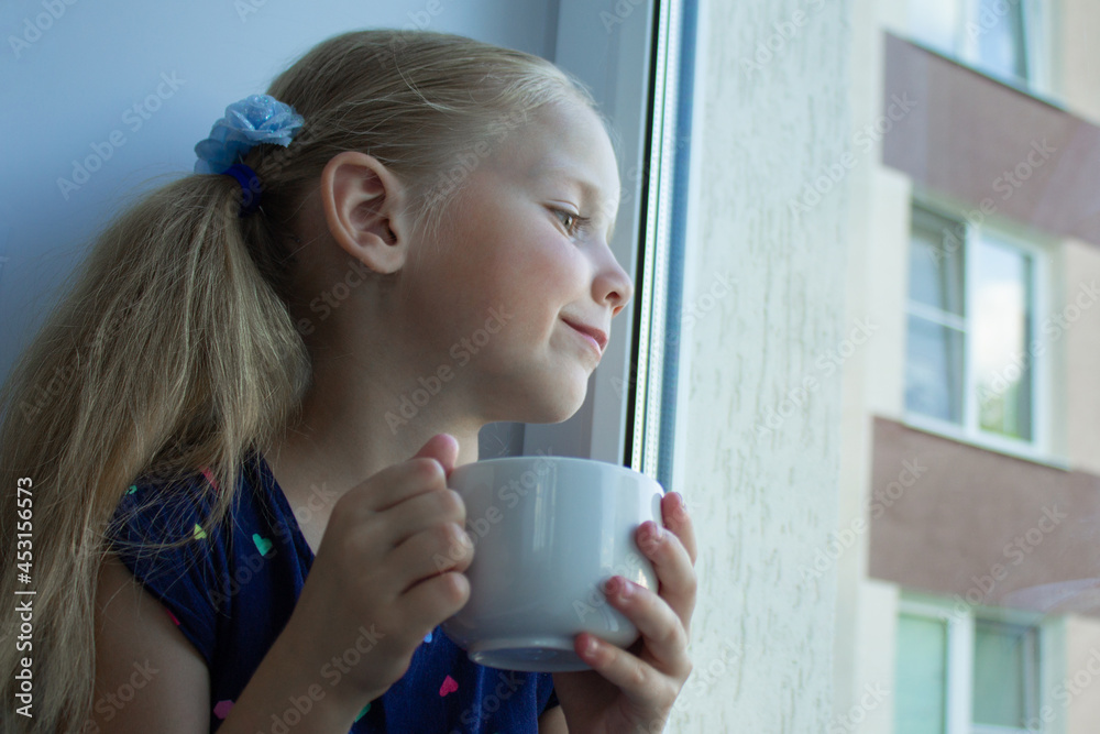 A beautiful blonde girl in a dark blue dress sits at home alone in quarantine in the summer on the windowsill by the window with a white cup of tea and looks sadly out. Tea drinking at lock down. A beautiful blonde girl in a dark blue dress sits at home alone in quarantine in the summer on the windowsill by the window with a white cup of tea and looks sadly out. Tea drinking at lock down.