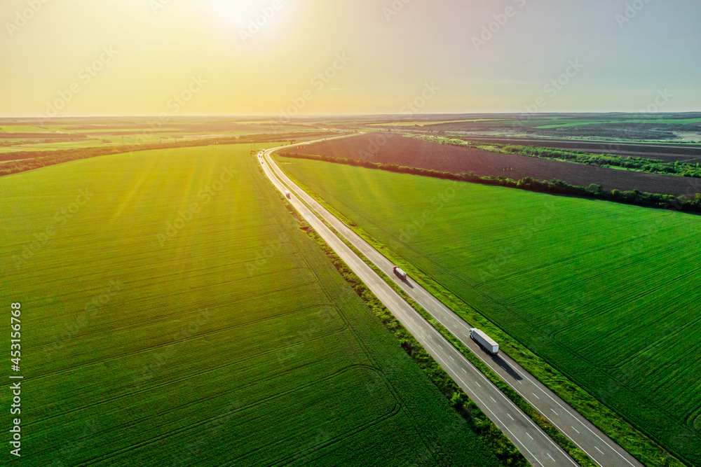 two white trucks driving on asphalt road along the green fields at sunset. seen from the air. Aerial view landscape. drone photography. cargo delivery