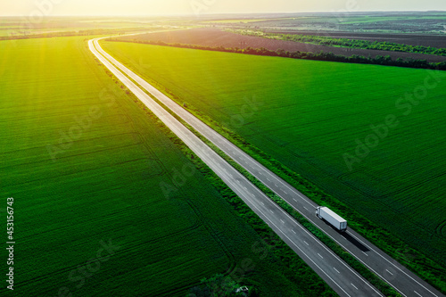Obraz na plátně white truck driving on asphalt road along the green fields at sunset