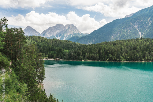 An emerald green Blindsee lake surrounded by forest and mountains, with hiking path on a side