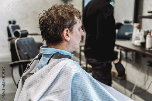 Visit to the barbershop.Young stylish man,businessman makes fashionable haircut.Barber, hairdresser,stylist and client,customer sitting in dark men's beauty salon with black chair covered with a cape © velirina