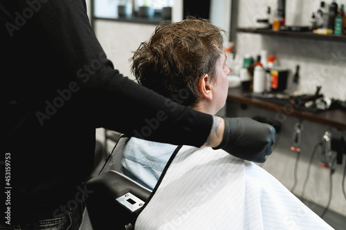 Visit to the barbershop.Young stylish man,businessman makes fashionable haircut.Barber, hairdresser,stylist and client,customer sitting in dark men's beauty salon with black chair covered with a cape