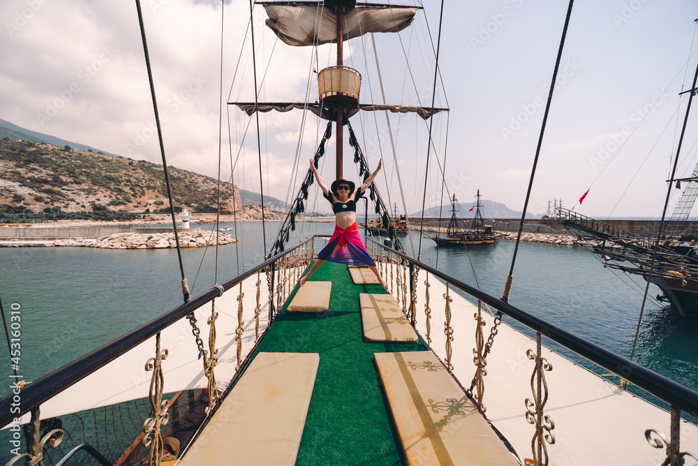Happy girl posing at old pirate ship on the water of Mediteranean sea. Tourist entertainment, coastal tour. Summer sunny day. Mountain shore of Alanya. Turkey.