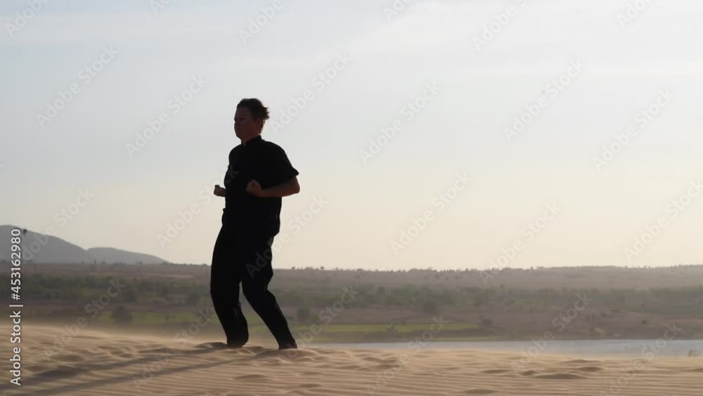 Woman in black is engaged in kung fu in the dunes at sunset. a lot of sand, golden sunset light. sports training. Silhouette of sport woman on the sunset, strong wind sweeps sand. close up plan
