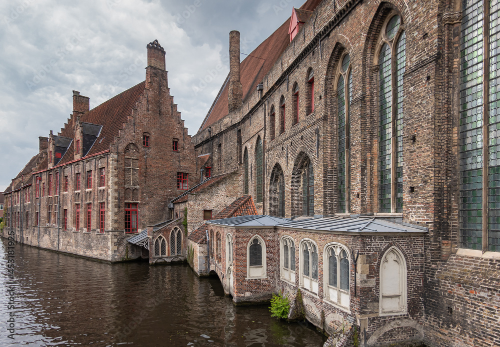 Brugge, Flanders, Belgium - August 4, 2021: Brown brick stone side facades of old Sint Jans Hospital now museum along black water canal at Mariabrug. Gray cloudscape.