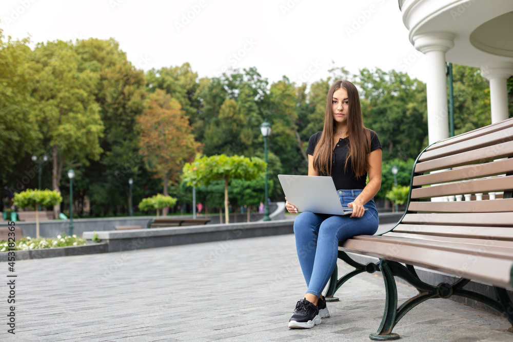 Education, business, technology, and Internet concept — Smiling European dark-haired girl in a black T-shirt and blue jeans sitting on a bench in the park in the summer and holding a laptop computer. Education, business, technology, and Internet concept — Smiling European dark-haired girl in a black T-shirt and blue jeans sitting on a bench in the park in the summer and holding a laptop computer.