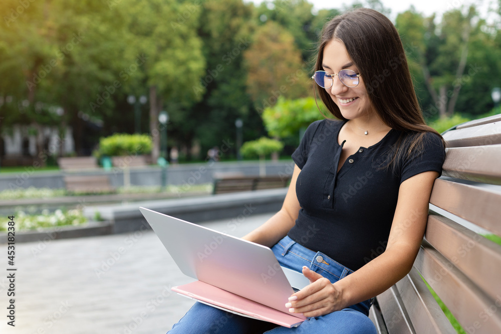 Obraz premium Education, technology, and Internet concept — A smiling European dark-haired student girl in modern clothes, sitting on a park bench in the summer and looking at her laptop with notebooks.
