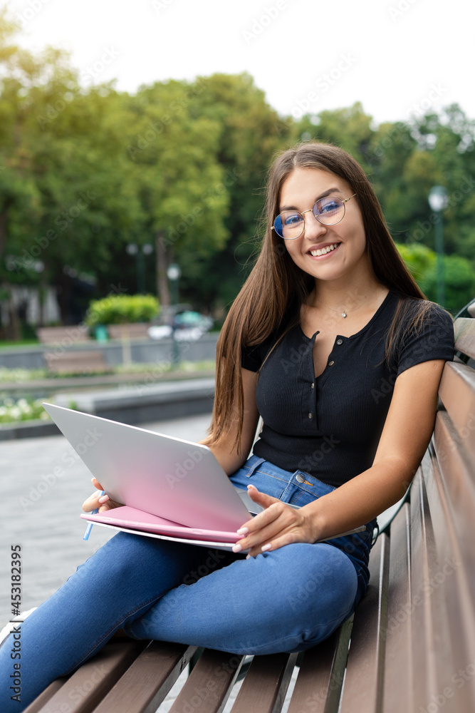 Obraz premium Freelance, business, technology, and Internet concept — A smiling European dark-haired women sitting on a park bench in the summer with a laptop, pen, and notebooks looking into the camera.
