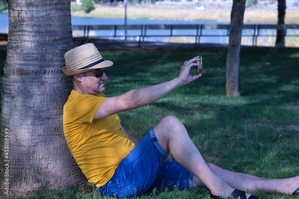 Mature adult Hispanic male wearing a hat and sunglasses, sitting on the grass and leaning against a tree taking a photo with his cell phone.