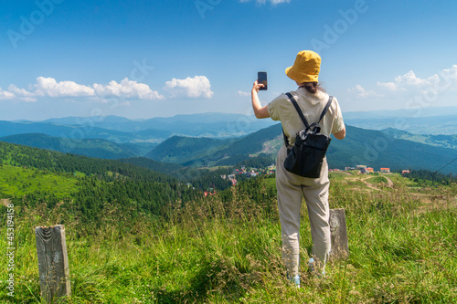 Traveler in a hat, resting in nature. A tourist girl photographs a panorama of the mountains by a mobile phone. Mountain beautiful landsc