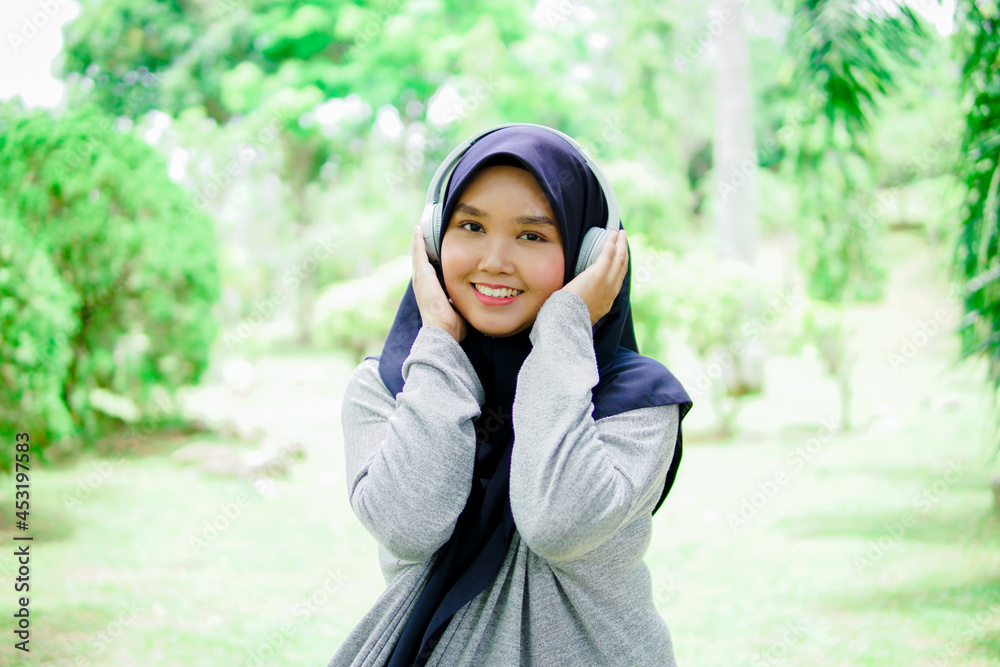Portrait of an attractive and beautiful young Asian Muslim woman listening to music with headphones in a park Portrait of an attractive and beautiful young Asian Muslim woman listening to music with headphones in a park