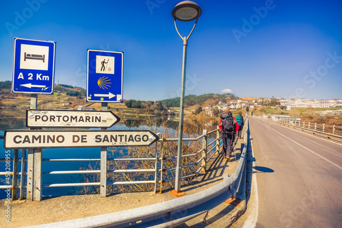 Pilgrims Hiker Backpackers Crossing Bridge over Lake Water to Portomarin, Spain, along the Way of St James Pilgrimage Trail Camino de Santiago