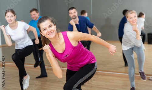 Positive young woman trainer doing aerobics exercises with group of adult people in dance center