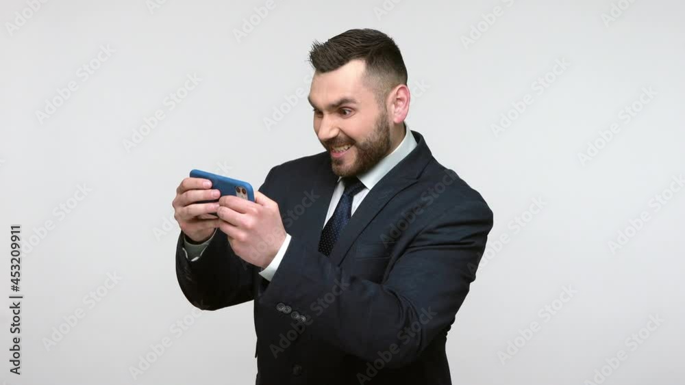 Bearded businessman in black official style suit playing virtual games on smart phone during break, winning, clenching fist, gadget addiction. Indoor studio shot isolated on gray background. Bearded businessman in black official style suit playing virtual games on smart phone during break, winning, clenching fist, gadget addiction. Indoor studio shot isolated on gray background.