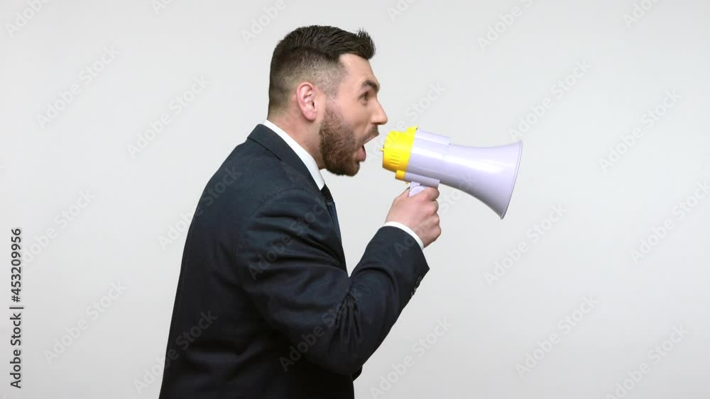 Side view portrait of young adult bearded businessman in black official style suit screaming at megaphone, making announcement, protest. Indoor studio shot isolated on gray background. Side view portrait of young adult bearded businessman in black official style suit screaming at megaphone, making announcement, protest. Indoor studio shot isolated on gray background.