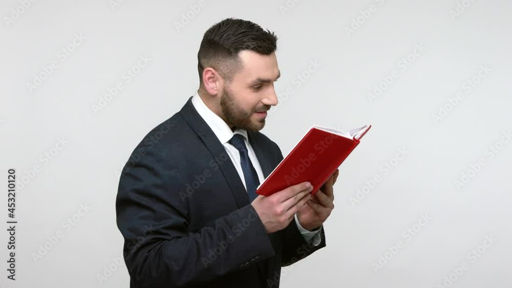 Portrait of good looking bearded businessman in black official style suit standing reading book, satisfied of plot, recommend for friends. Indoor studio shot isolated on gray background. Portrait of good looking bearded businessman in black official style suit standing reading book, satisfied of plot, recommend for friends. Indoor studio shot isolated on gray background.