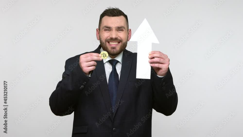 Portrait of bearded businessman in black official style suit holding white arrow and btc, showing growth of bitcoin, banking, deposit concept. Indoor studio shot isolated on gray background. Portrait of bearded businessman in black official style suit holding white arrow and btc, showing growth of bitcoin, banking, deposit concept. Indoor studio shot isolated on gray background.