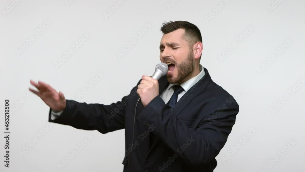 Portrait of positive cheerful bearded businessman in black official style suit standing with microphone in hands, singing songs in karaoke. Indoor studio shot isolated on gray background. Portrait of positive cheerful bearded businessman in black official style suit standing with microphone in hands, singing songs in karaoke. Indoor studio shot isolated on gray background.