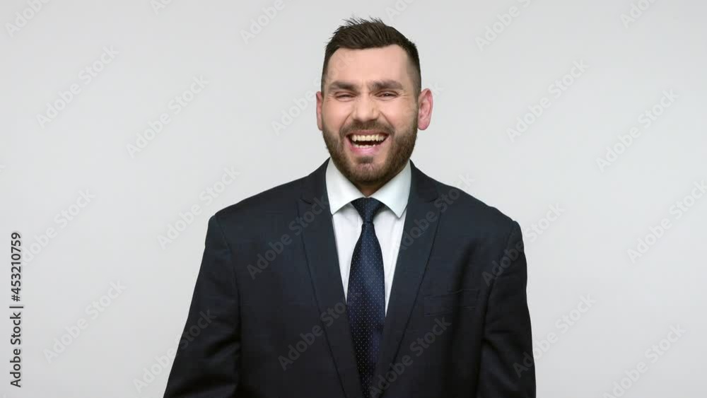 Optimistic smiling young adult bearded businessman in black official style suit laughing out loud, holding belly, watching humorous TV show. Indoor studio shot isolated on gray background. Optimistic smiling young adult bearded businessman in black official style suit laughing out loud, holding belly, watching humorous TV show. Indoor studio shot isolated on gray background.