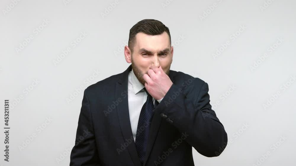 Shameful bearded businessman in black official style suit pinching nose, gesturing go away with your stinking breath, disgusting smell. Indoor studio shot isolated on gray background. Shameful bearded businessman in black official style suit pinching nose, gesturing go away with your stinking breath, disgusting smell. Indoor studio shot isolated on gray background.