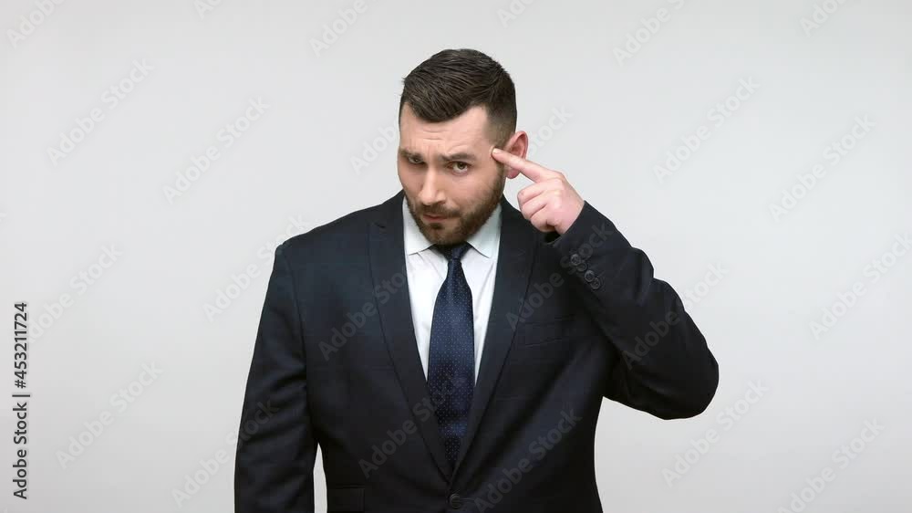 Upset bearded businessman in black official style suit twisting finger near temple, making stupid gesture and sadly looking at camera. Indoor studio shot isolated on gray background. Upset bearded businessman in black official style suit twisting finger near temple, making stupid gesture and sadly looking at camera. Indoor studio shot isolated on gray background.
