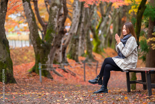 Asian woman tourist sitting on bench in the park red maple tree leaves falling down on footpath in autumn. Smiling female enjoy outdoor lifestyle activity travel vacation on season change in Japan