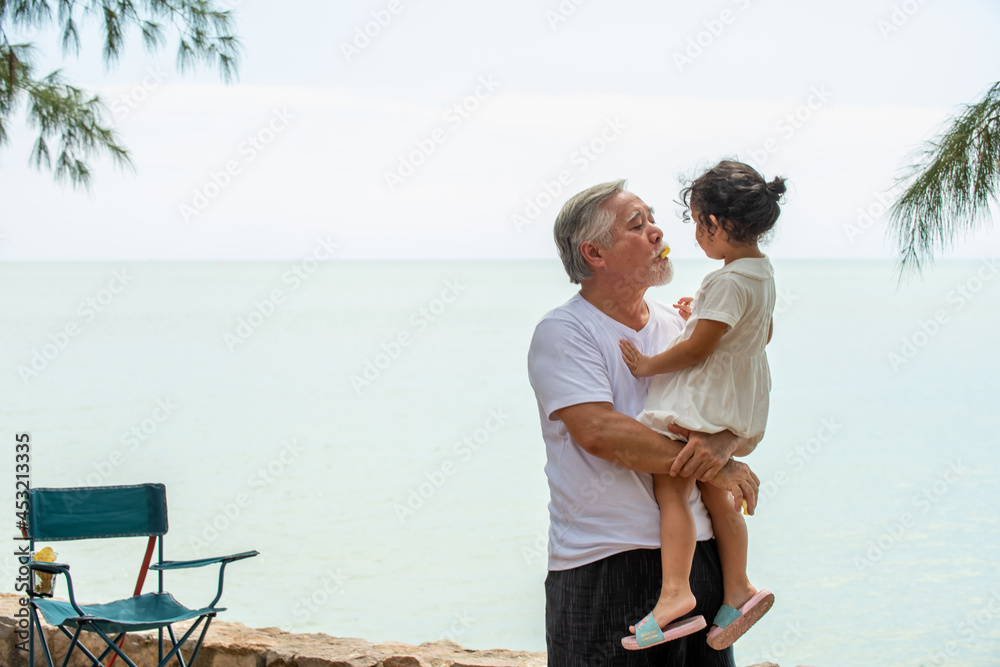 Happy Asian family on summer travel vacation. Grandfather carrying and playing with grandchild girl on the beach. Senior man with granddaughter enjoy and having fun outdoor activity lifestyle together