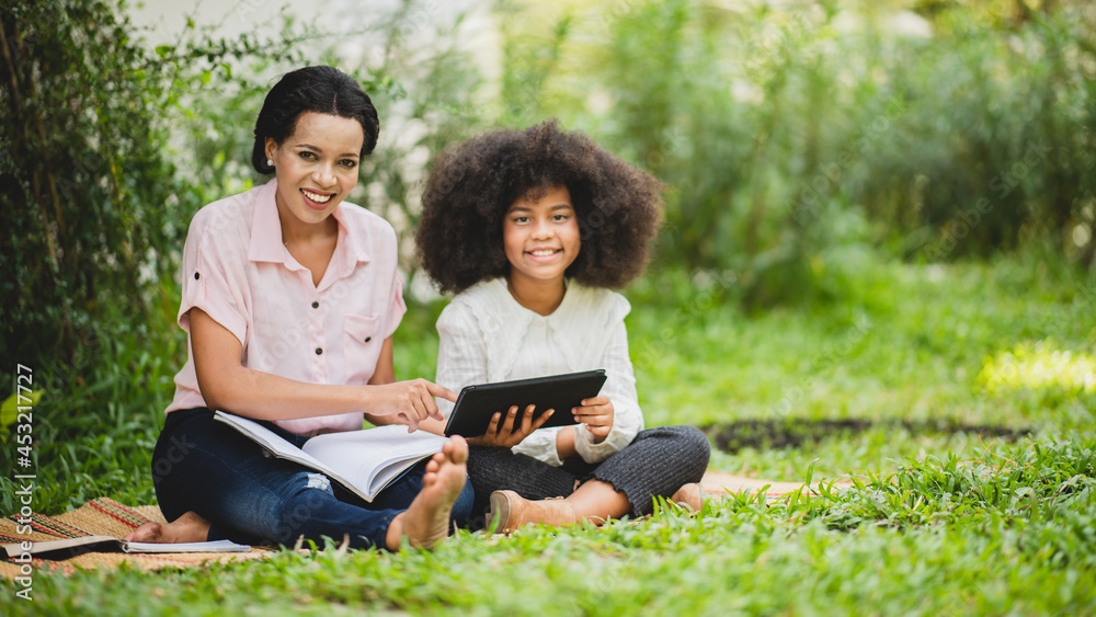 Cheerful young beautiful mother sitting with son with curly hair in park and helping him while using digital tablet while studying