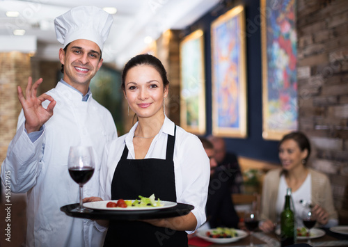 Portrait of positive young waitress with cook chef standing in restaurant