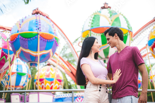 Handsome young man smiling and holding woman by waist standing in theme park while looking at girlfriend with love