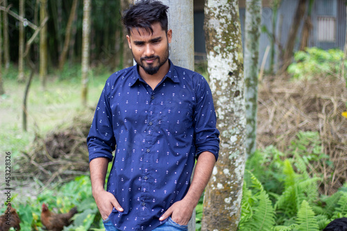 A young boy is wearing a dark blue shirt and standing with his hands in his pockets. His short beard and his behind blur green nature background.