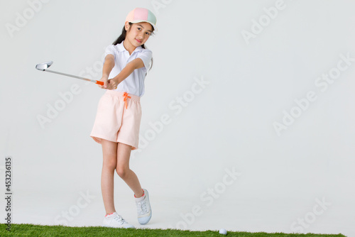 Portrait isolated studio shot of Asian little professional girl golfer in sport cloth uniform holding golf club driver in hands ready to drive and hit ball on green grass in front of white background