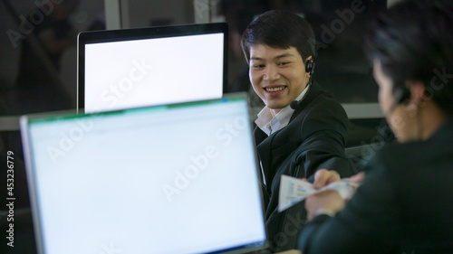 Asian smile man wearing headphone headset and working with happy face and sevice mind in call center in front of computer screen in office at night. Overtime, late work and 24 hours service concept photo