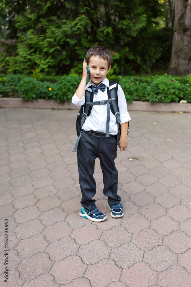 a beautiful little boy in a white shirt and gray trousers a schoolboy with a bow around his neck is standing in the school yard with a backpack on his shoulders and a phone in his hands