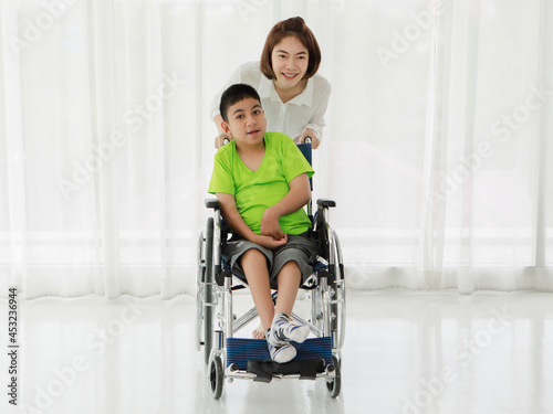 Smiling Asian nurse rolling smiling ethnic boy with disability sitting in wheelchair photo