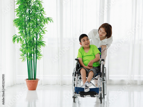 Smiling Asian nurse rolling smiling ethnic boy with disability sitting in wheelchair