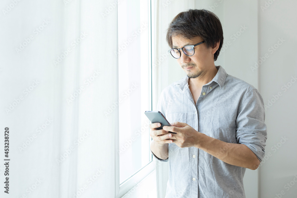 Asian businessman texting on smartphone standing next to window.