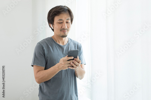 Asian guy using voice assistant on cellphone with earpod by the window at home.