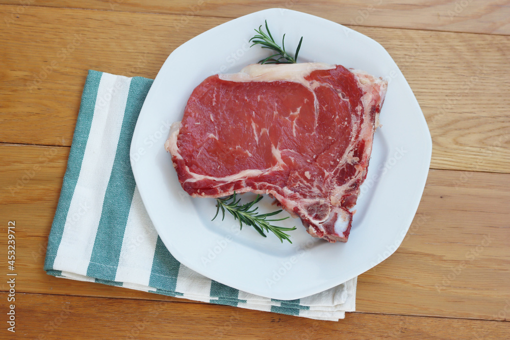 Raw beef meat steak on a white plate on wooden table with fresh rosemary herbs ready to cook Raw beef meat steak on a white plate on wooden table with fresh rosemary herbs ready to cook