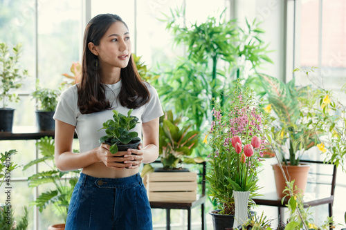 Girl long hair wear t shirt holding red Dieffenbachia tree pot smiling standing in white mirror room looking at camera. Surround by air filtering plants colorful flower spring sunlight feeling warm.