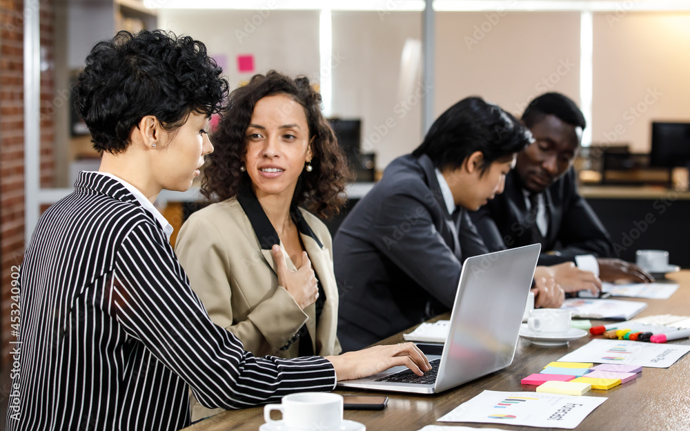 Close up to two mixed race caucasian african beautiful smart professional businesswomen talking, discussing job strategy, using laptop for working in indoor office with background of male colleagues. Close up to two mixed race caucasian african beautiful smart professional businesswomen talking, discussing job strategy, using laptop for working in indoor office with background of male colleagues.