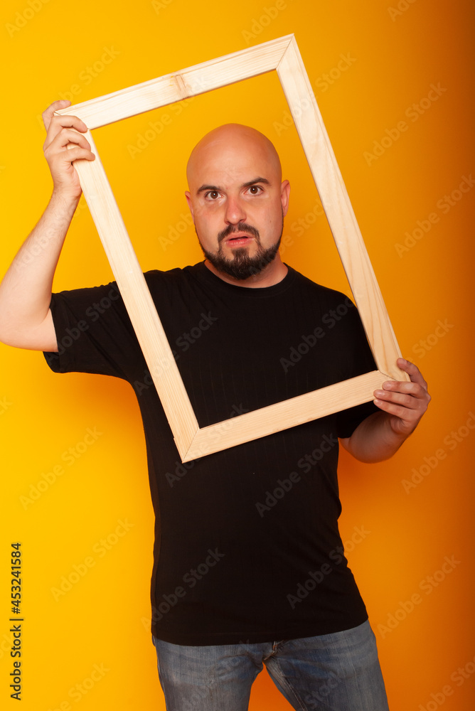 a man with a bald head without hair of European appearance holds a frame by his face looking into the camera on a yellow background