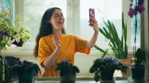 Pretty happy young woman talking on video call on the cellphone sitting near a window. Home interior with natural plants. Real time. Internet communication concept. photo