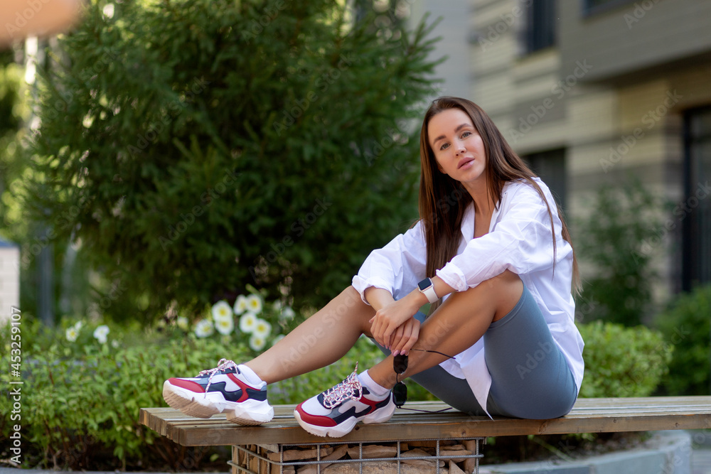 Naklejka premium Fashion portrait of a young fashionable woman, dressed in sports clothes, sitting on the bench, enjoys rest in a city park, summer street fashion. portrait of a joyful woman