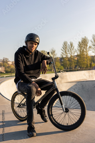 Teenage boy wearing helmet sits on black bmx, low bike on ramp in spring, man doing sports, outdoor activity photo