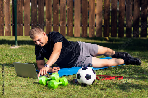 Outdoor portrait of the young man in a sports uniform is resting, stretching on the lawn , watches a movie and studies from a laptop , a social network in garden