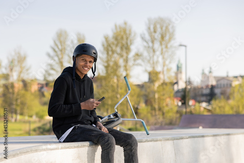 A student sits on a ramp in a park near the city. The boy is smiling sitting with his helmet unzipped, his bike, a bmx, lies next to him.
