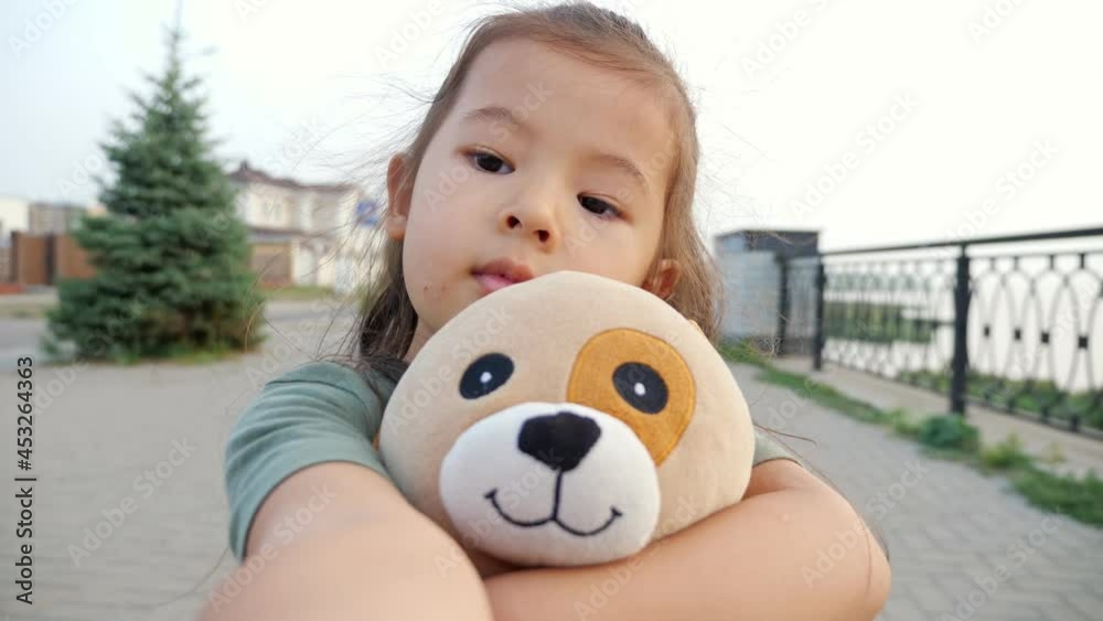 Pretty little Asian girl with soft toy dog makes selfie standing on city embankment with railing and fir tree in summer evening slow motion Pretty little Asian girl with soft toy dog makes selfie standing on city embankment with railing and fir tree in summer evening slow motion